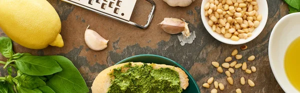 Top view of baguette slice with pesto sauce on plate near fresh ingredients and cooking utensils on stone surface, panoramic shot — Stock Photo