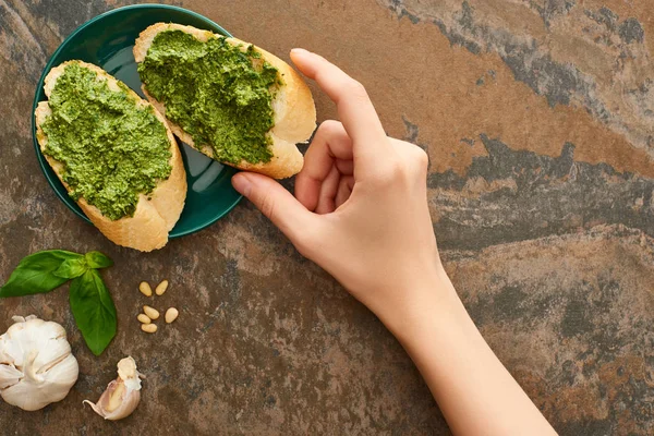 Vista cortada da mulher tomando fatias de baguete com molho pesto da placa perto de ingredientes frescos na superfície de pedra — Fotografia de Stock