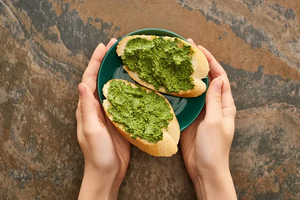 Vista recortada de la mujer sosteniendo el plato con rebanadas de baguette y deliciosa salsa de pesto en la superficie de piedra - foto de stock