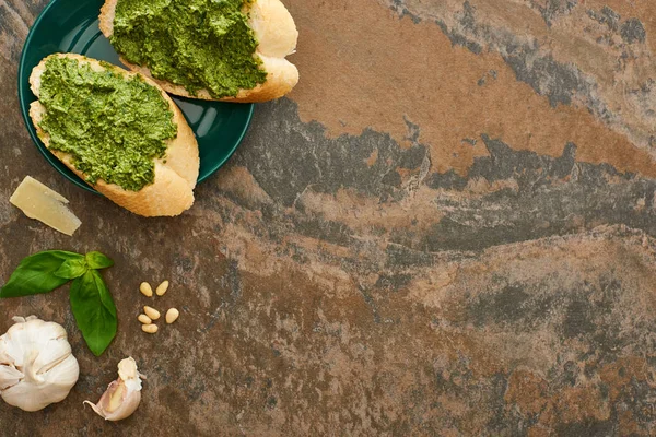 Vue de dessus des tranches de baguette avec sauce pesto sur l'assiette près des ingrédients frais sur la surface de la pierre — Photo de stock