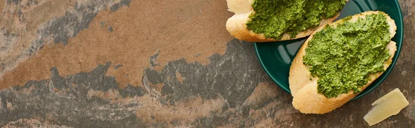 Top view of baguette slices with pesto sauce on plate near parmesan on stone surface, panoramic shot — Stock Photo