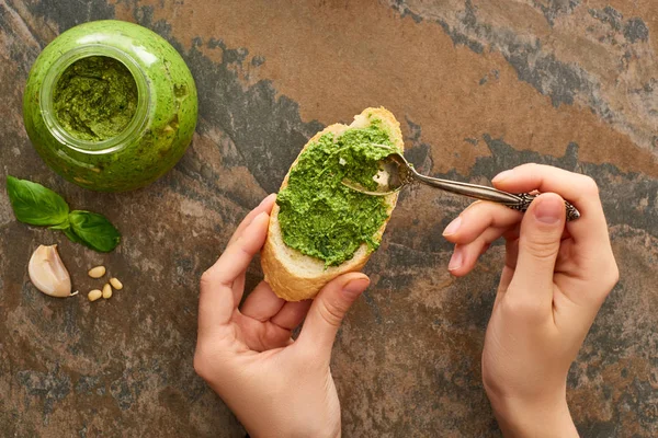 Vista cortada da mulher adicionando molho pesto com colher em fatia de baguete na superfície de pedra — Fotografia de Stock