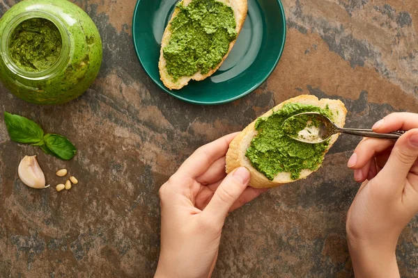 Vista recortada de la mujer añadiendo salsa de pesto con cuchara en la rebanada de baguette en la superficie de piedra - foto de stock