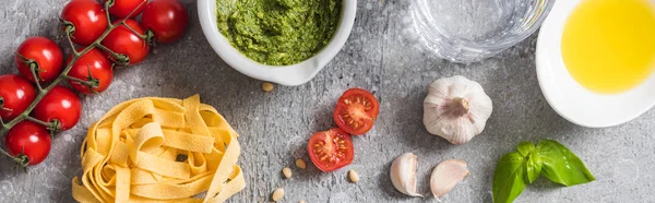 Top view of raw Pappardelle near tomatoes, garlic, basil, pine nuts, olive oil, water and pesto sauce on grey surface, panoramic shot — Stock Photo