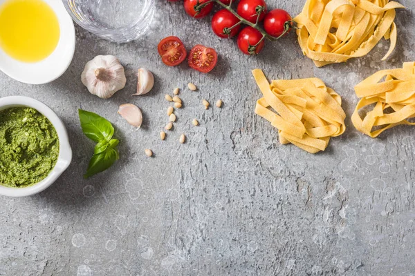 Vista superior de Pappardelle crudo cerca de tomates, ajo, albahaca, piñones, aceite de oliva, agua y salsa de pesto en la superficie gris - foto de stock