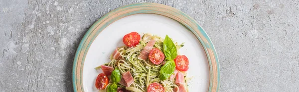 Top view of cooked Pappardelle with tomatoes, basil and prosciutto on plate on grey surface, panoramic shot — Stock Photo