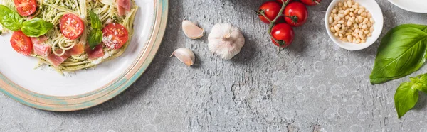 Top view of cooked Pappardelle with tomatoes, basil and prosciutto near ingredients on grey surface, panoramic shot — Stock Photo