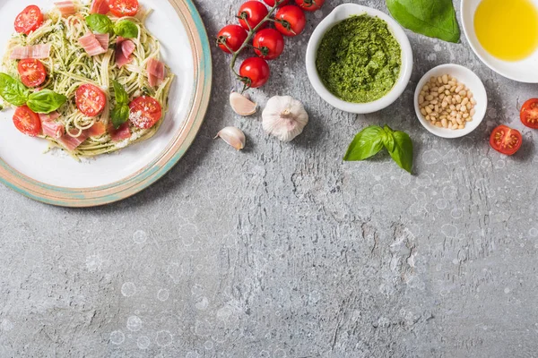 Blick von oben auf gekochte Pappardelle mit Tomaten, Basilikum und Schinken in der Nähe frischer Zutaten auf grauer Oberfläche — Stockfoto