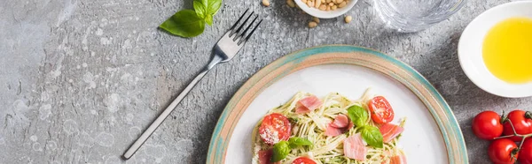 Top view of cooked Pappardelle with tomatoes, basil and prosciutto near ingredients and fork on grey surface, panoramic shot — Stock Photo