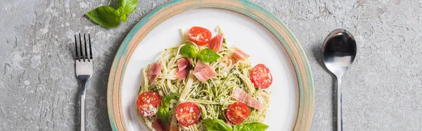 Top view of cooked Pappardelle with tomatoes, basil and prosciutto near cutlery on grey surface, panoramic shot — Stock Photo