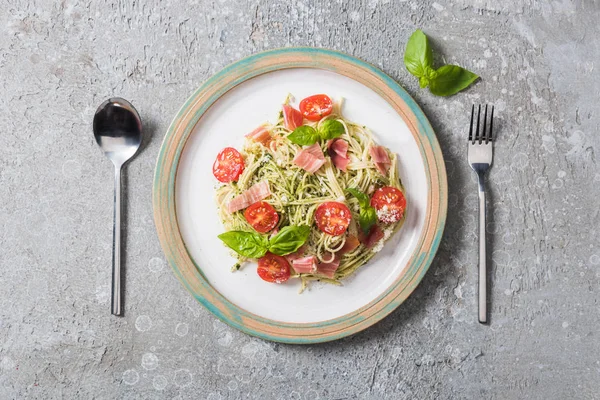 Top view of cooked Pappardelle with tomatoes, basil and prosciutto near cutlery on grey surface — Stock Photo