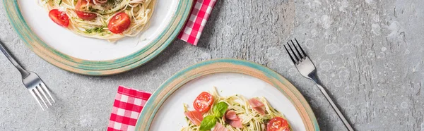 Top view of Pappardelle with tomatoes, basil and prosciutto on plates on plaid napkins with forks on grey surface, panoramic shot — Stock Photo