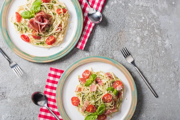 Pappardelle mit Tomaten, Basilikum und Prosciutto auf Tellern auf karierten Servietten mit Besteck auf grauer Oberfläche — Stockfoto