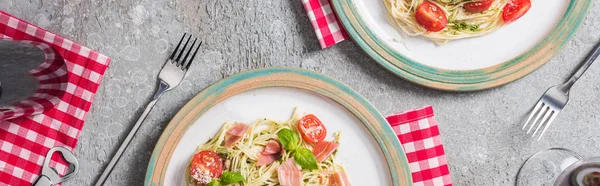 Top view of Pappardelle with tomatoes, basil and prosciutto on plates on plaid napkins with forks near red wine on grey surface, panoramic shot — Stock Photo