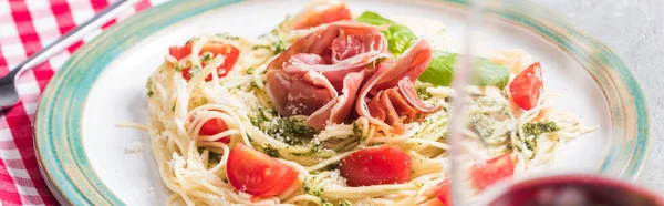 Selective focus of red wine and Pappardelle with tomatoes, basil and prosciutto on grey surface with plaid napkin, panoramic shot — Stock Photo