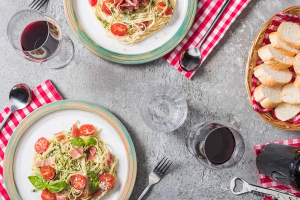 Vista superior de Pappardelle servido con tomates, albahaca y prosciutto en servilletas a cuadros con cubiertos cerca de baguette, vino tinto y agua en superficie gris — Stock Photo