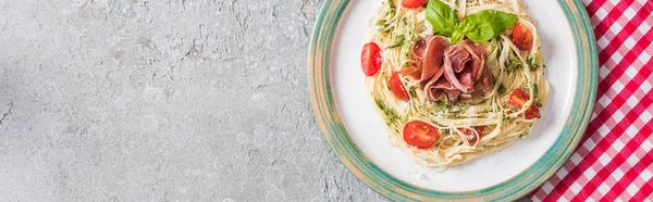 Vue du dessus de la Pappardelle servie avec tomates, basilic et prosciutto sur serviette à carreaux sur surface grise, vue panoramique — Photo de stock