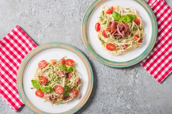 Draufsicht auf servierte Pappardelle mit Tomaten, Basilikum und Prosciutto auf karierten Servietten auf grauer Oberfläche — Stockfoto