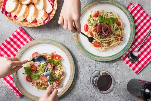 Vista ritagliata delle donne che mangiano pappardelle con pomodori, basilico e prosciutto vicino baguette, vino rosso sulla superficie grigia — Foto stock