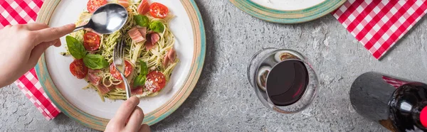 Cropped view of woman eating Pappardelle with tomatoes, basil and prosciutto near red wine on grey surface, panoramic shot — Stock Photo