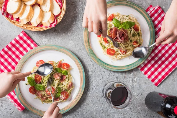 Cropped view of women eating Pappardelle with tomatoes, basil and prosciutto near baguette, red wine on grey surface — Stock Photo
