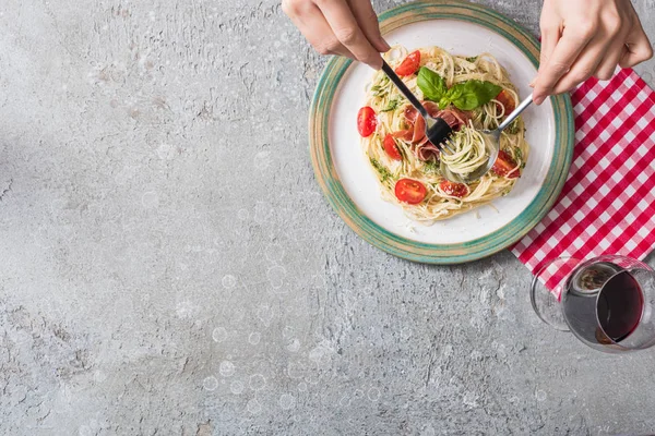 Partial view of woman eating Pappardelle with tomatoes, basil and prosciutto on plaid napkin near glass of red wine on grey surface — Stock Photo