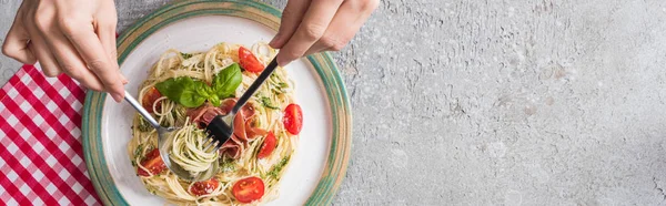 Teilansicht einer Frau, die Pappardelle mit Tomaten, Basilikum und Prosciutto auf karierter Serviette auf grauer Oberfläche isst, Panoramaaufnahme — Stockfoto