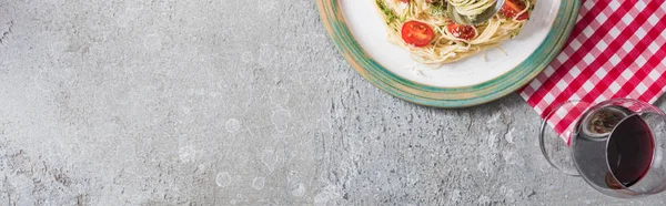 Vista dall'alto di Pappardelle con pomodori, basilico e prosciutto su tovagliolo plaid vicino a calice di vino rosso su superficie grigia, foto panoramica — Foto stock