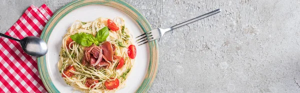 Top view of served Pappardelle with tomatoes, basil and prosciutto on plaid napkin with cutlery on grey surface, panoramic shot — Stock Photo