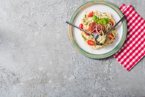 Vista superior de Pappardelle servido com tomates, manjericão e prosciutto em guardanapo xadrez com talheres na superfície cinza — Fotografia de Stock