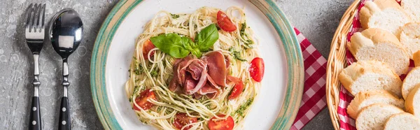 Top view of served Pappardelle with tomatoes, basil and prosciutto near baguette and cutlery on grey surface, panoramic shot — Stock Photo