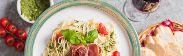 Selective focus of served Pappardelle with tomatoes, basil and prosciutto near baguette, red wine, pesto, tomatoes on grey surface, panoramic shot — Stock Photo