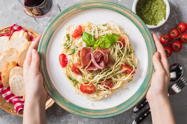Selective focus of woman holding plate with Pappardelle with tomatoes, basil and prosciutto near baguette, red wine, pesto, tomatoes and cutlery on grey surface — Stock Photo