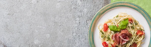 Vue du dessus de la Pappardelle cuite aux tomates, basilic et prosciutto sur assiette sur serviette verte sur surface grise, vue panoramique — Photo de stock