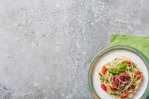 Top view of cooked Pappardelle with tomatoes, basil and prosciutto on plate on green napkin on grey surface — Stock Photo