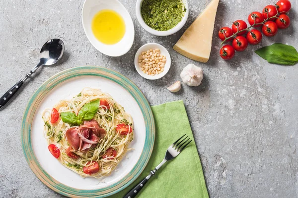 Top view of cooked Pappardelle with tomatoes, basil and prosciutto near ingredients and cutlery on grey surface — Stock Photo