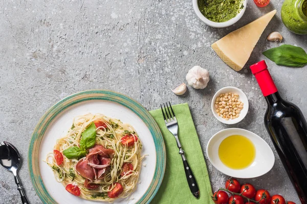 Top view of cooked Pappardelle with tomatoes, basil and prosciutto near ingredients, red wine and cutlery on grey surface — Stock Photo