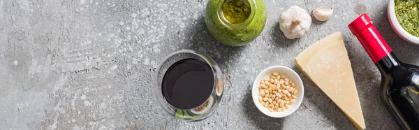 Vue du dessus du parmesan, des pignons de pin, de l'ail, de la sauce pesto et du vin rouge sur la surface grise, vue panoramique — Photo de stock