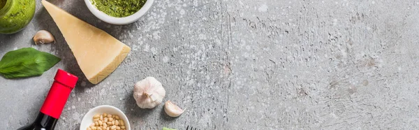 Top view of Parmesan, pine nuts, garlic, pesto sauce and bottle of red wine on grey surface, panoramic shot — Stock Photo