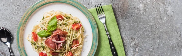 Top view of cooked Pappardelle with tomatoes, basil and prosciutto on plate on green napkin with cutlery on grey surface, panoramic shot — Stock Photo