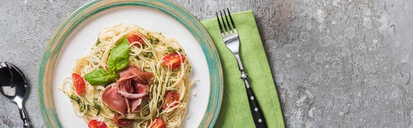 Vista superior de Pappardelle cocido con tomates, albahaca y prosciutto en plato en servilleta verde con cubiertos en superficie gris, plano panorámico — Stock Photo