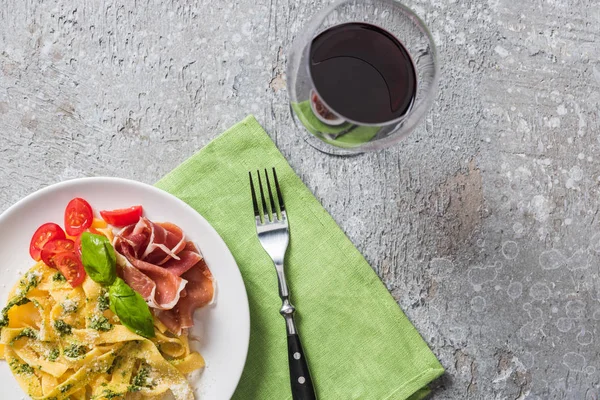 Vue du dessus de la Pappardelle cuite aux tomates, basilic et prosciutto près du vin rouge à la surface grise — Photo de stock