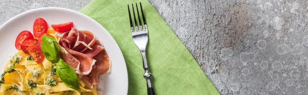 Top view of cooked Pappardelle with tomatoes, basil and prosciutto near fork on grey surface, panoramic shot — Stock Photo