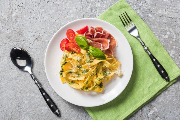 Top view of cooked Pappardelle with tomatoes, basil and prosciutto near cutlery on grey surface — Stock Photo