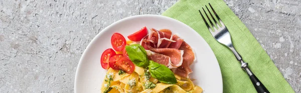 Top view of cooked Pappardelle with tomatoes, basil and prosciutto near fork on grey surface, panoramic shot — Stock Photo