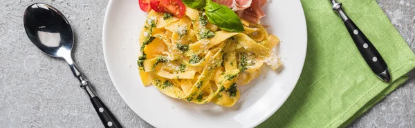 Top view of cooked Pappardelle with tomatoes, basil and prosciutto near cutlery on grey surface, panoramic shot — Stock Photo