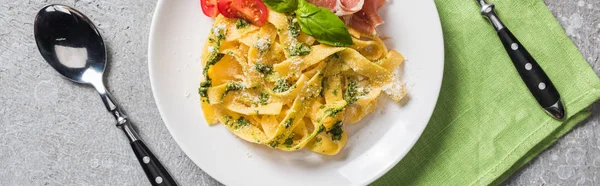 Top view of cooked Pappardelle with tomatoes, basil and prosciutto near cutlery on grey surface, panoramic shot — Stock Photo