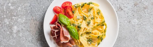 Top view of cooked Pappardelle with tomatoes, pesto sauce and prosciutto on grey surface, panoramic shot — Stock Photo