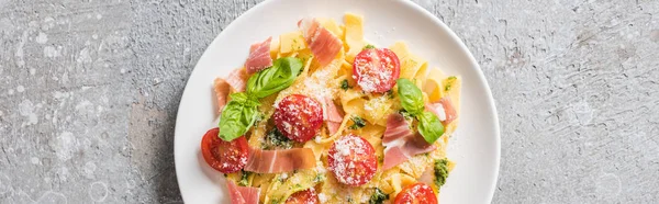 Top view of cooked Pappardelle with tomatoes, pesto sauce and prosciutto on grey surface, panoramic shot — Stock Photo