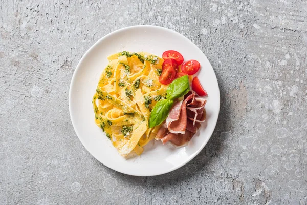 Top view of cooked Pappardelle with tomatoes, pesto sauce and prosciutto on grey surface — Stock Photo